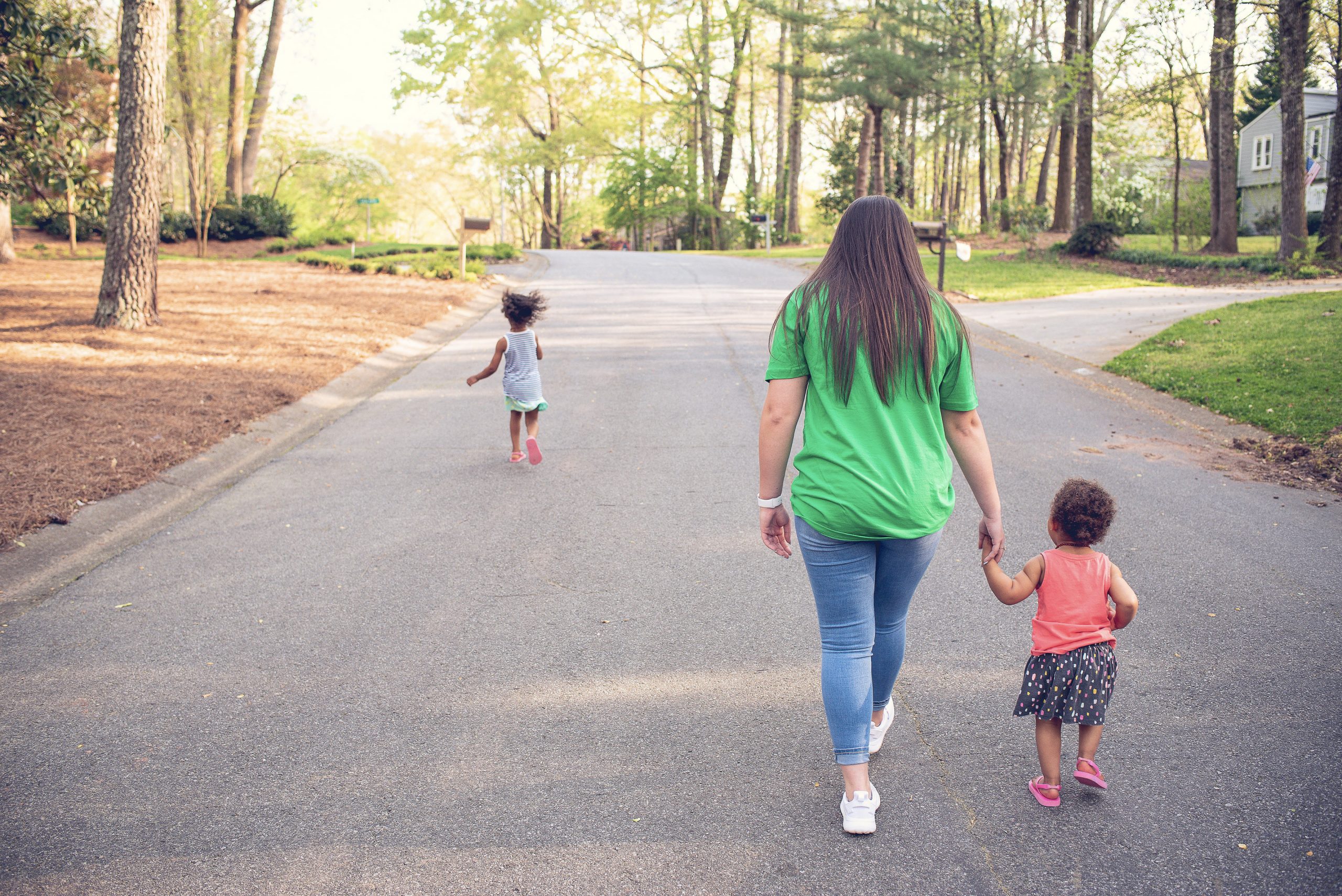 A babysitter in a green SitterTree t-shirt holds hands with a young child as they walk down a quiet, tree-lined neighborhood street, while another child runs ahead in the warm, sunlit setting: Find a nanny near me