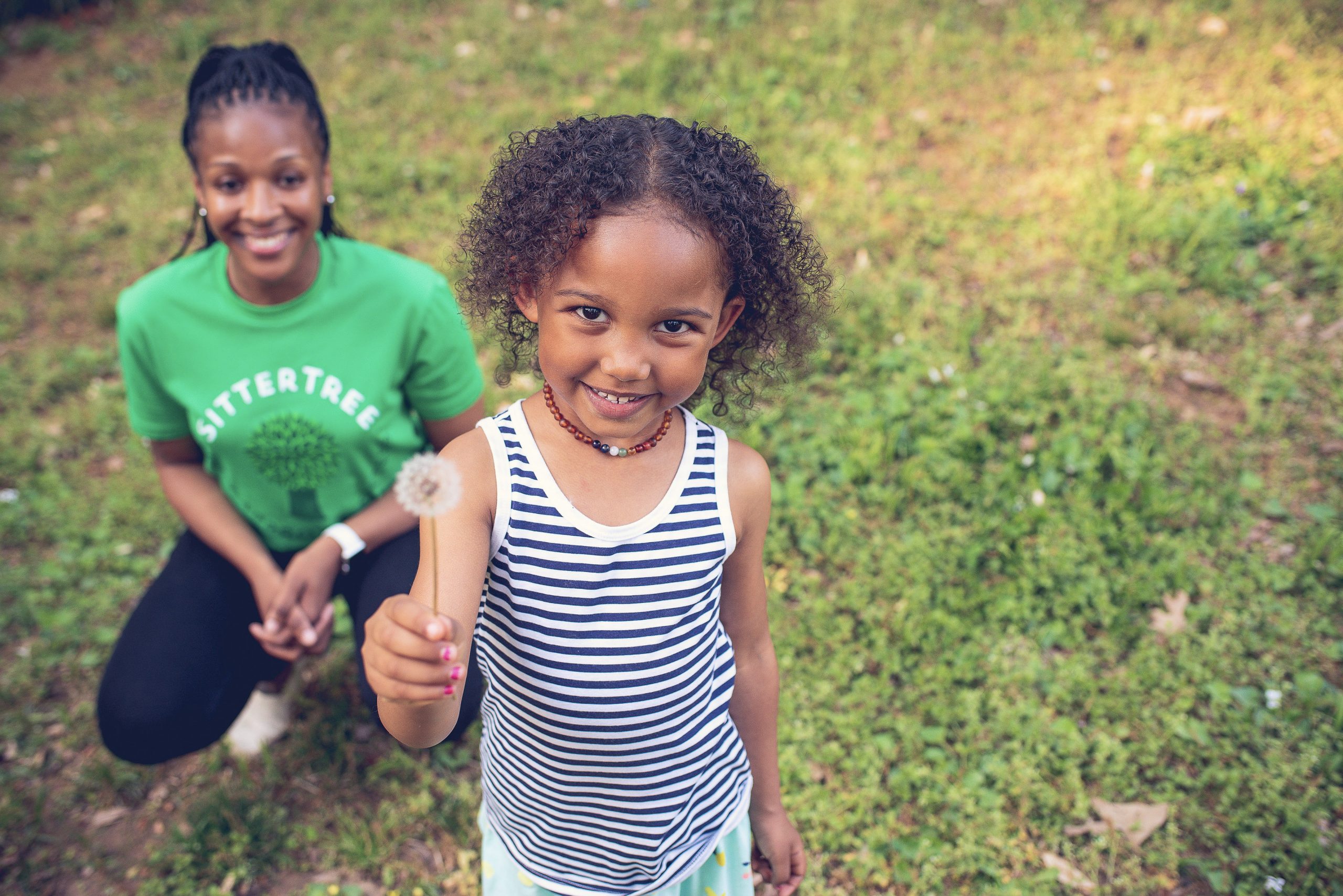 Young girl smiling and holding a dandelion in an outdoor grassy setting, with a babysitter in a green SitterTree t-shirt crouching behind her, creating a playful and cheerful moment: how to verify babysitter references