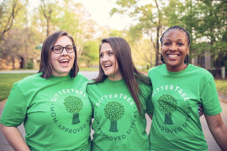 Three cheerful babysitters wearing matching green SitterTree t-shirts, standing outdoors in a sunlit, leafy neighborhood, smiling and enjoying each other's company: Find a nanny near me