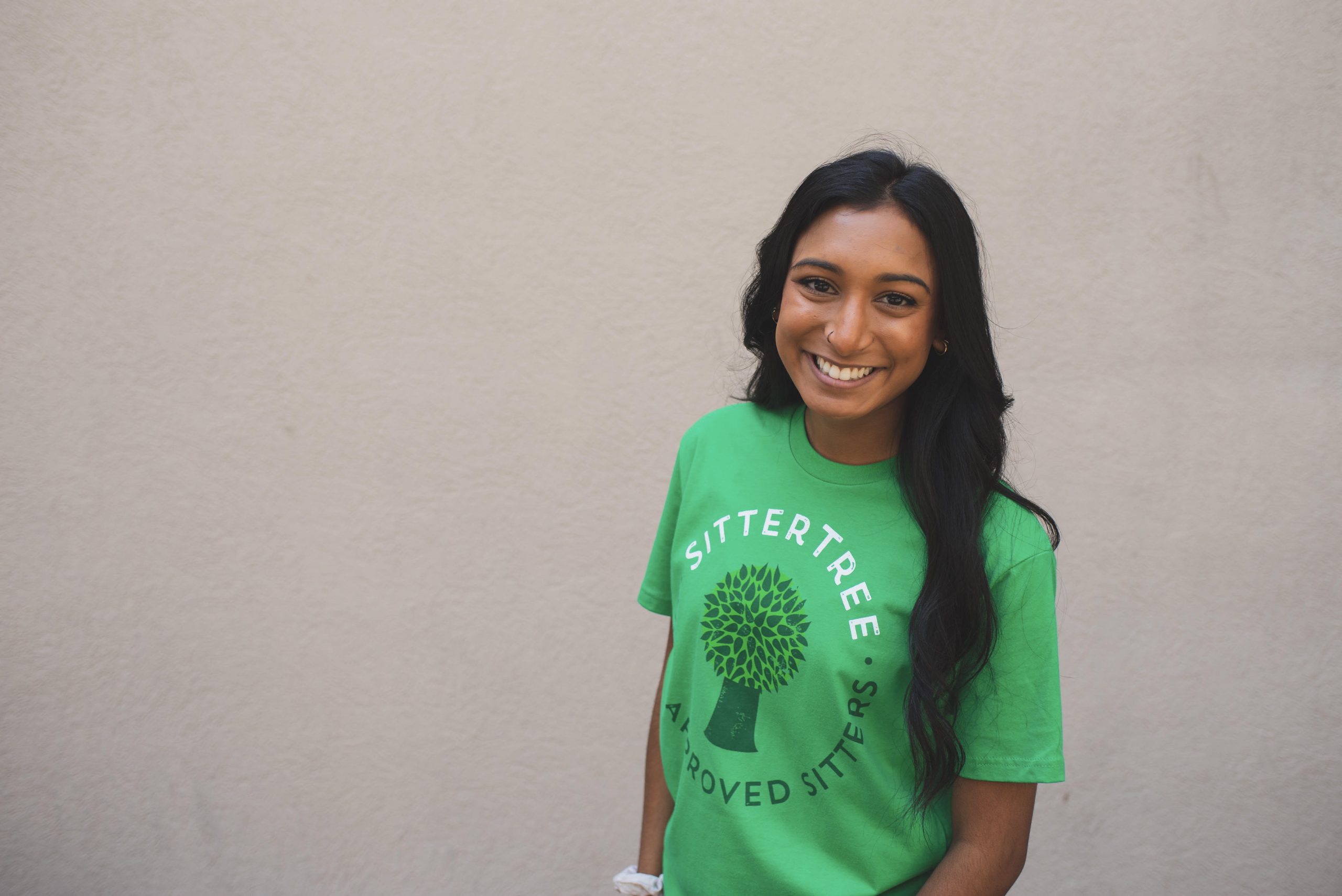 A smiling babysitter wearing a green SitterTree t-shirt standing against a neutral beige background, radiating warmth and friendliness: How to find a nanny