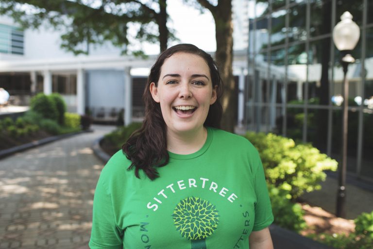 "Smiling babysitter wearing a green SitterTree t-shirt, standing outdoors in a landscaped area with trees and modern buildings in the background: how to verify babysitter references