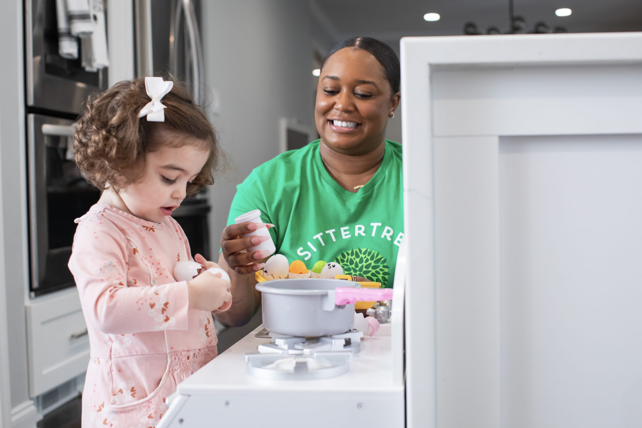 Babysitter in a green SitterTree t-shirt smiling and playing pretend kitchen with a young girl wearing a pink dress and a white bow, engaging in imaginative play together in a modern home setting: creative babysitting activities