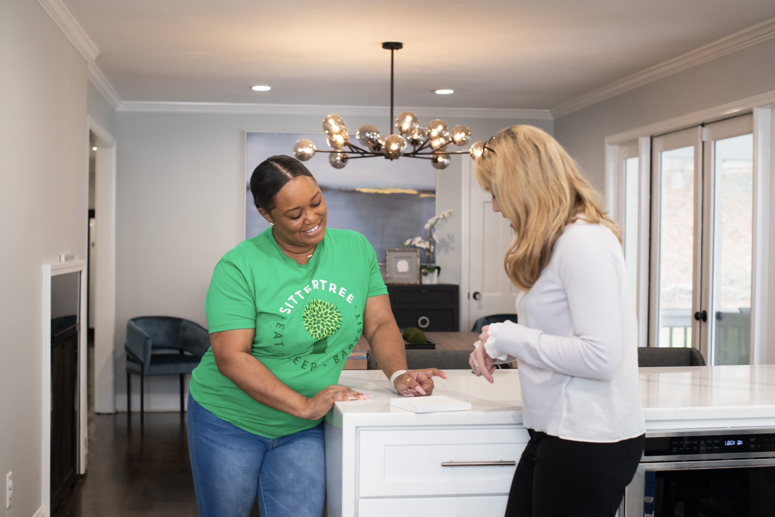 Babysitter in a green SitterTree t-shirt smiling and talking with a parent in a bright, modern kitchen, fostering a friendly and professional interaction: how to handle feedback from parents