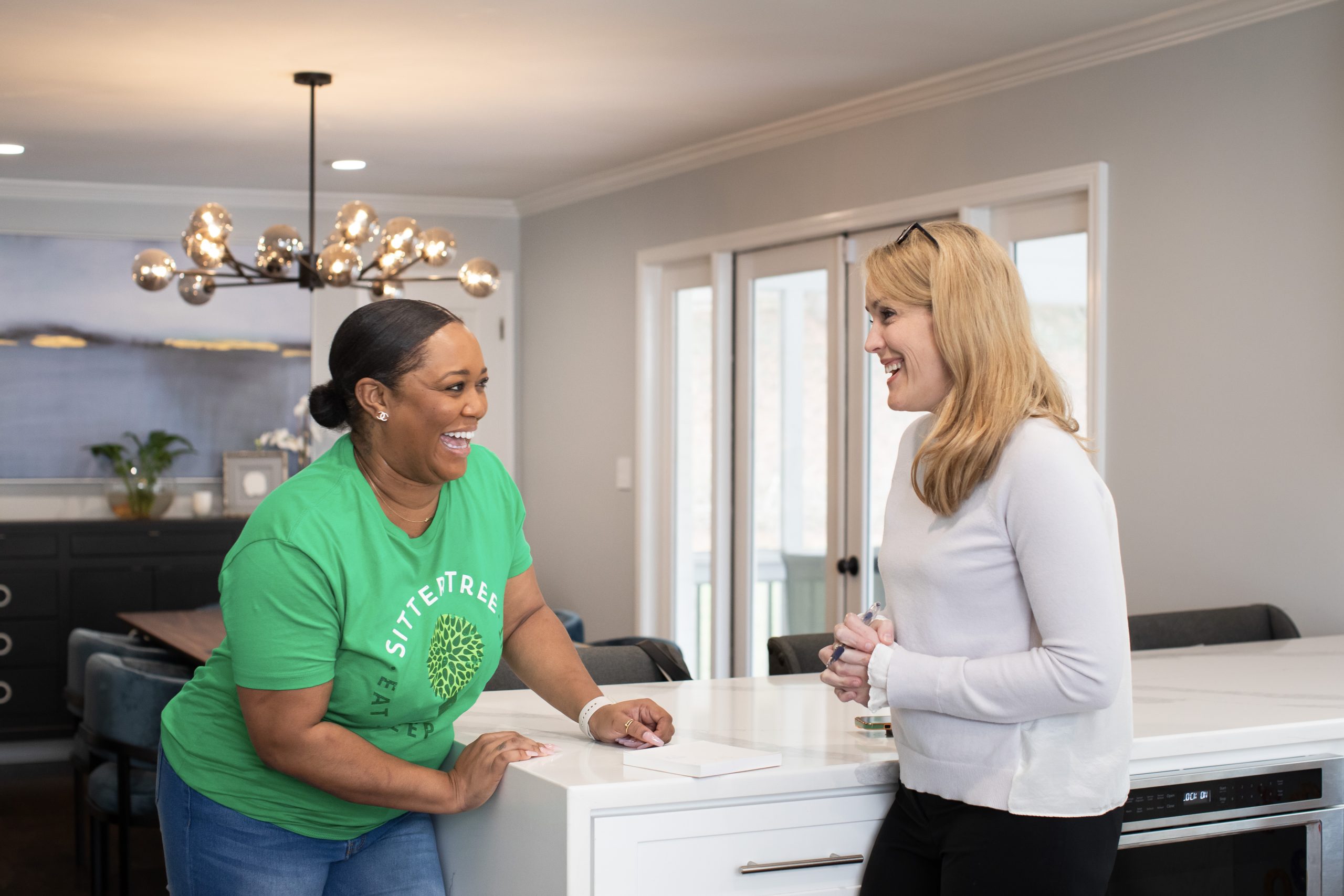 Babysitter in a green SitterTree t-shirt laughing and chatting with a parent in a bright and modern kitchen, creating a warm and friendly atmosphere: how to maintain open communication with your babysitter