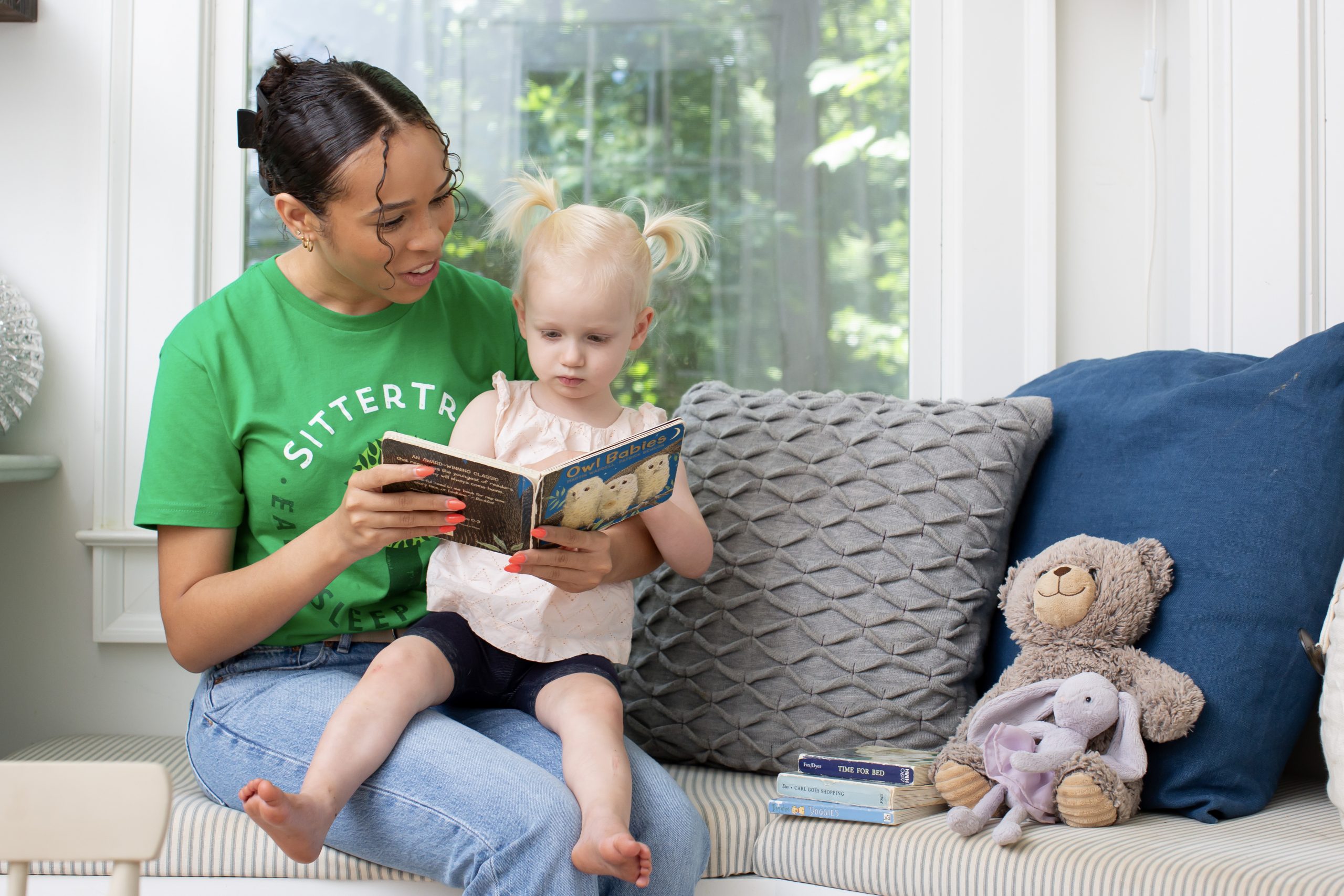 "A babysitter in a green SitterTree t-shirt sitting on a cushioned bench, reading a children's book to a young girl with blonde pigtails, surrounded by cozy pillows and stuffed animals in a bright and inviting room: How to find a nanny