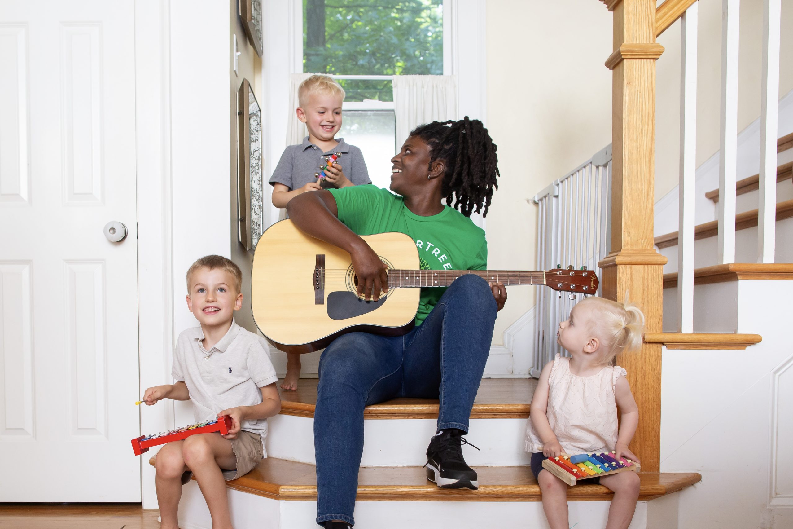 Babysitter in a green SitterTree t-shirt playing a guitar while sitting on stairs with three young children holding musical instruments, creating a joyful and interactive music session: how to address performance issues with your babysitter