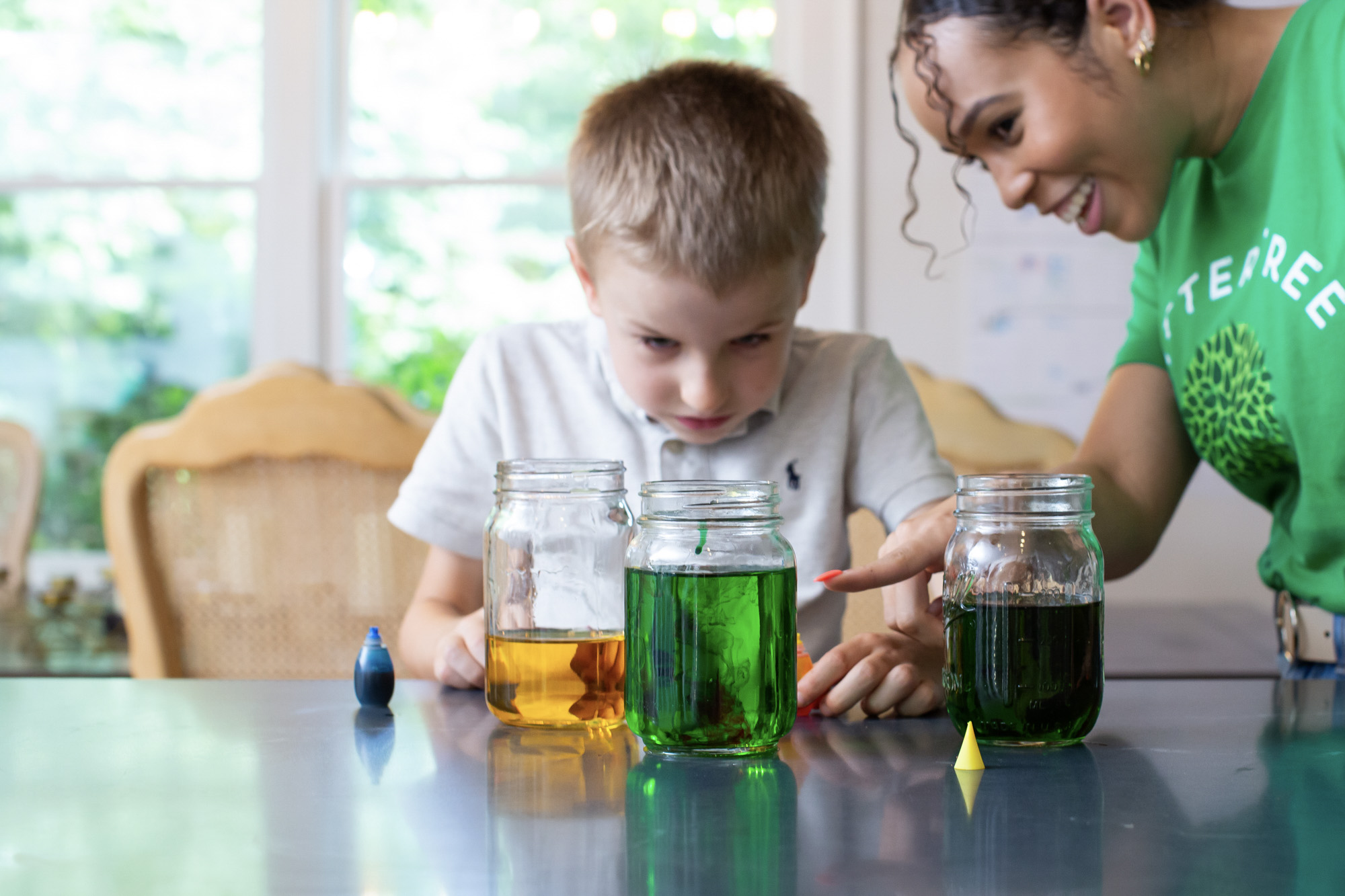 Babysitter in a green SitterTree t-shirt smiling and guiding a young boy as they conduct a fun science experiment with jars of colorful liquids, fostering curiosity and hands-on learning: fun babysitting games