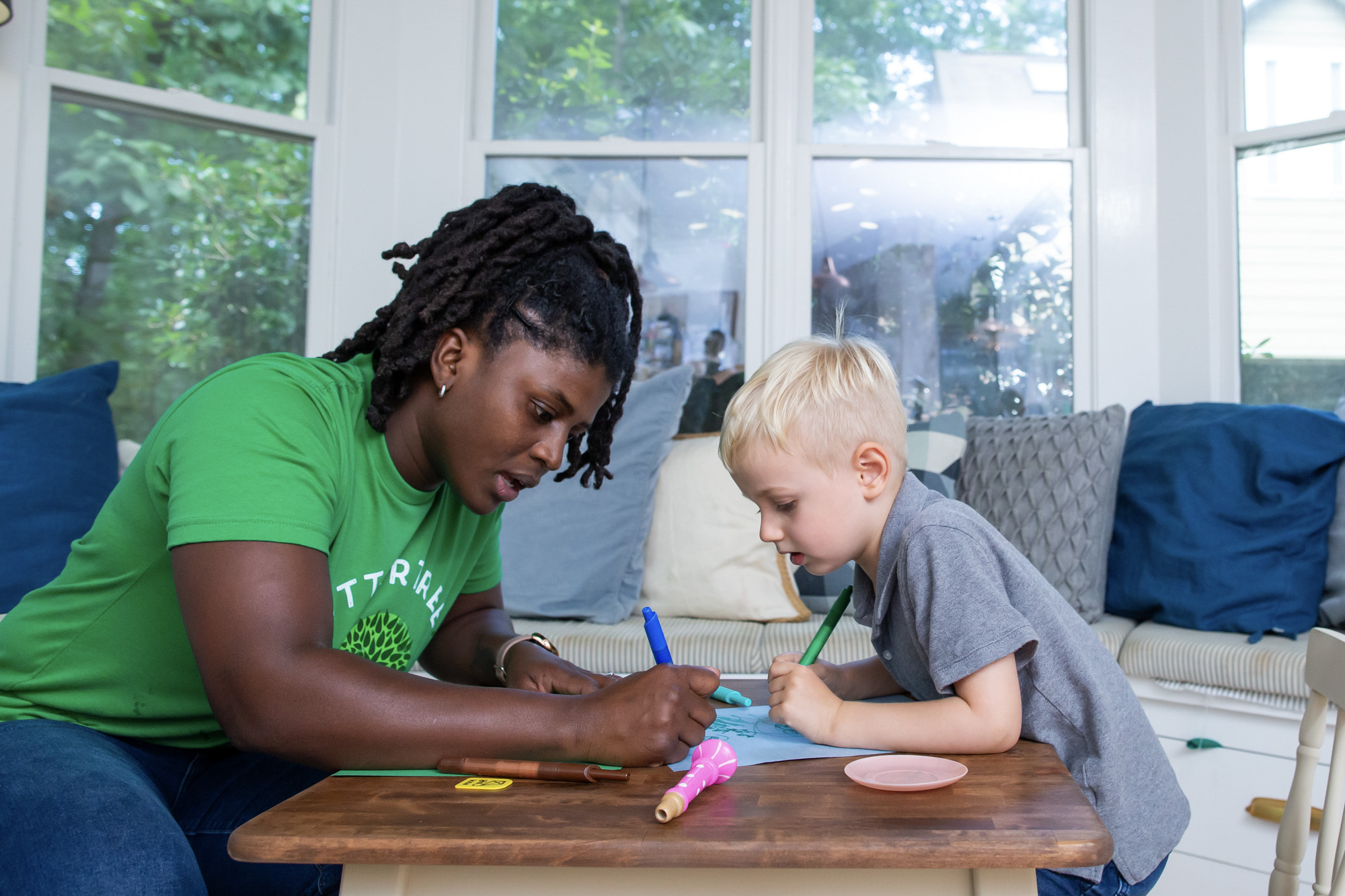 Babysitter in a green SitterTree t-shirt sitting at a small table with a young boy, both focused on drawing and coloring together in a bright, cozy room with large windows and cushions in the background: How to handle tantrums as a babysitter