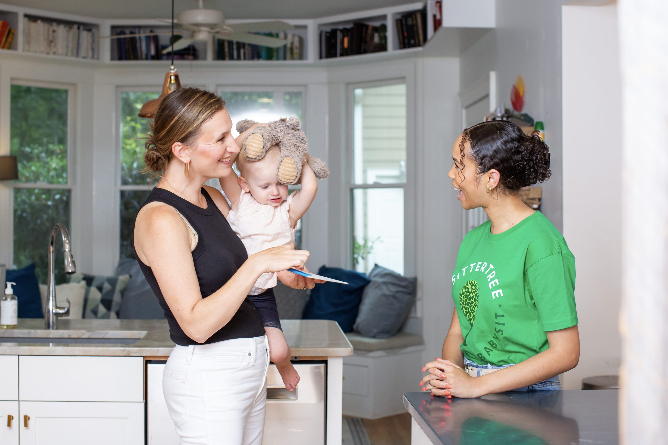 Babysitter in a green SitterTree t-shirt chatting with a smiling parent holding a toddler who is playfully wearing a stuffed animal on their head, in a bright and cozy kitchen setting: how to show appreciation to your babysitter