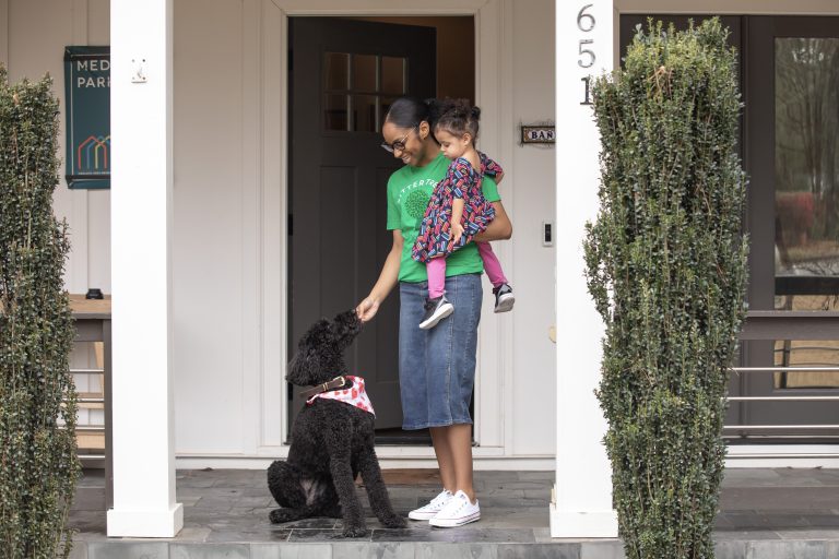 A babysitter wearing a green SitterTree t-shirt stands on a porch holding a toddler in colorful clothing, smiling as they interact with a black poodle wearing a bandana: How to find a babysitter near me