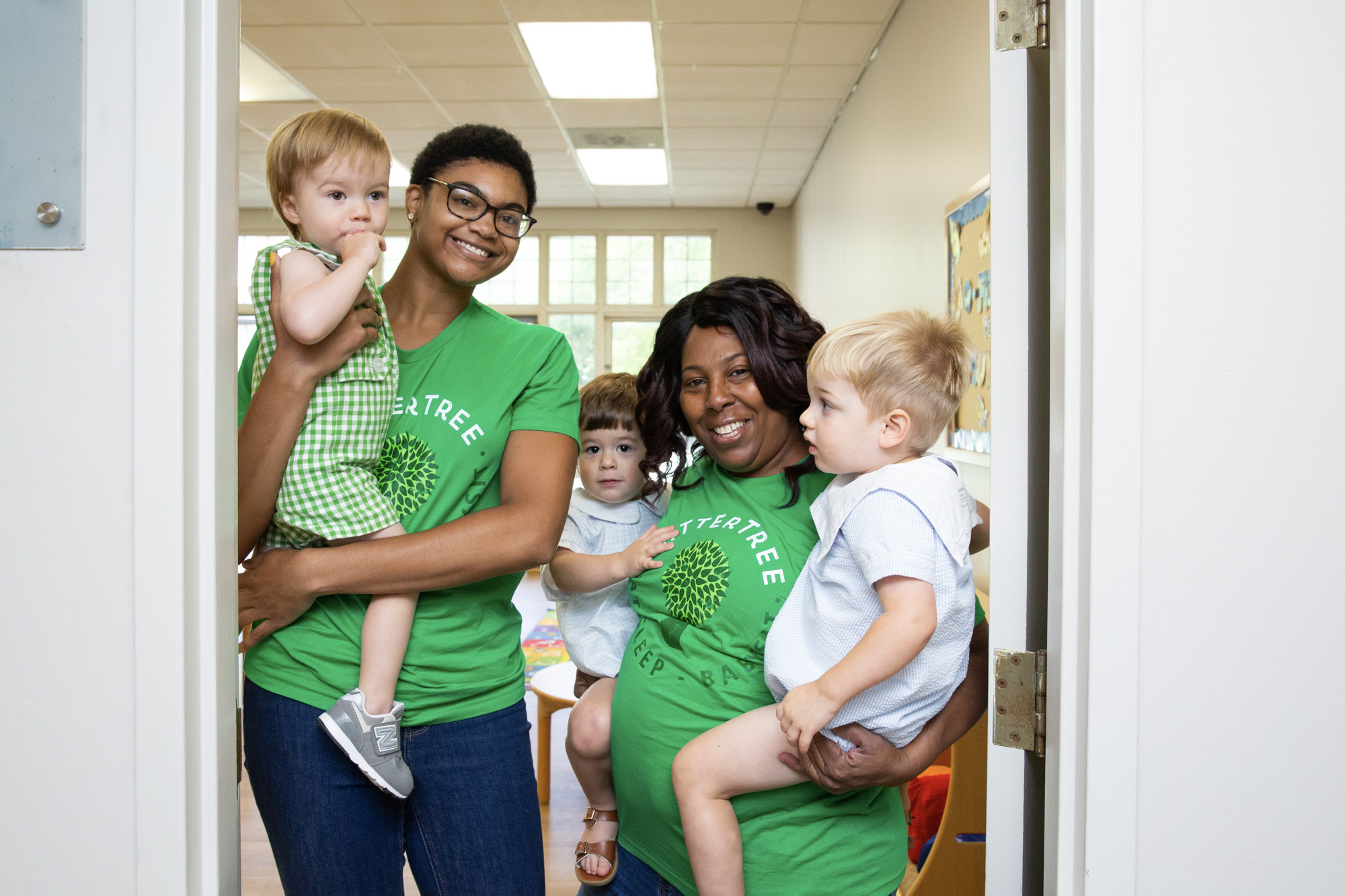 Two babysitters wearing green SitterTree t-shirts smile while holding toddlers in their arms, standing in a bright classroom doorway with a bulletin board in the background: Interview questions for childcare teachers