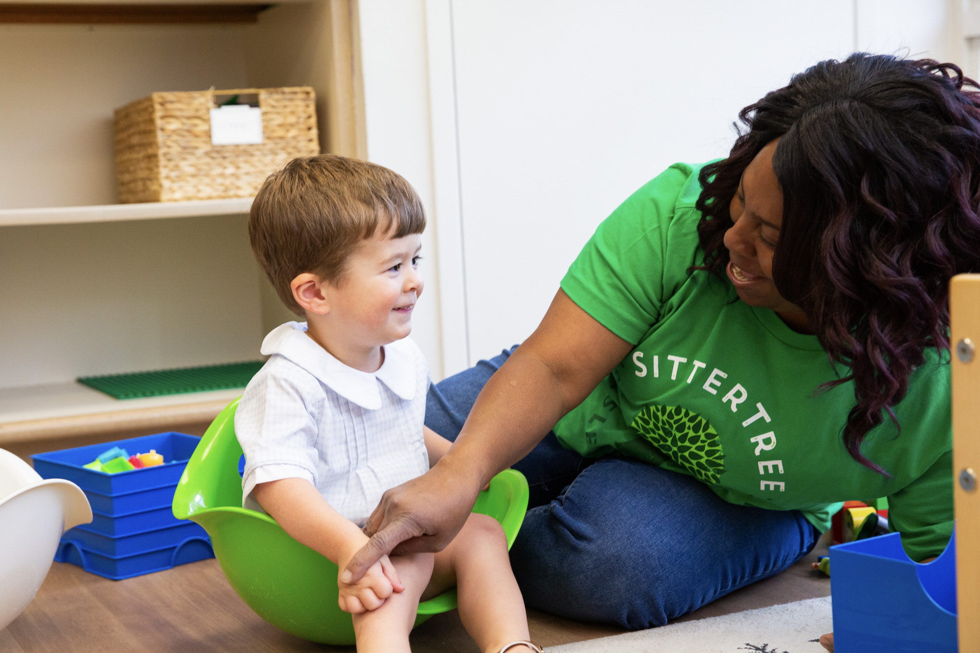 Babysitter in a green SitterTree t-shirt smiling warmly at a young child sitting in a green toy seat, engaging in playful interaction in a classroom setting with toys and storage bins in the background: skills every babysitter should develop