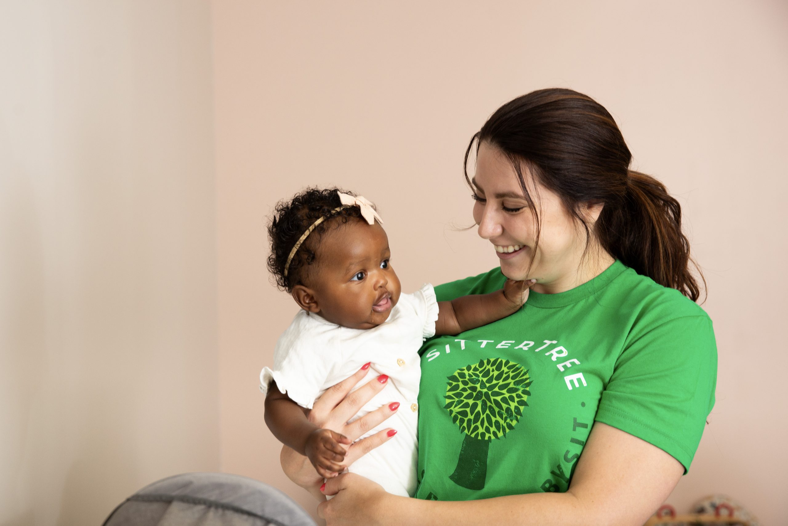 Babysitter in a green SitterTree t-shirt smiling warmly while holding an adorable baby wearing a white outfit and a bow headband, creating a loving and nurturing moment: what to expect on the first day with a new babysitter