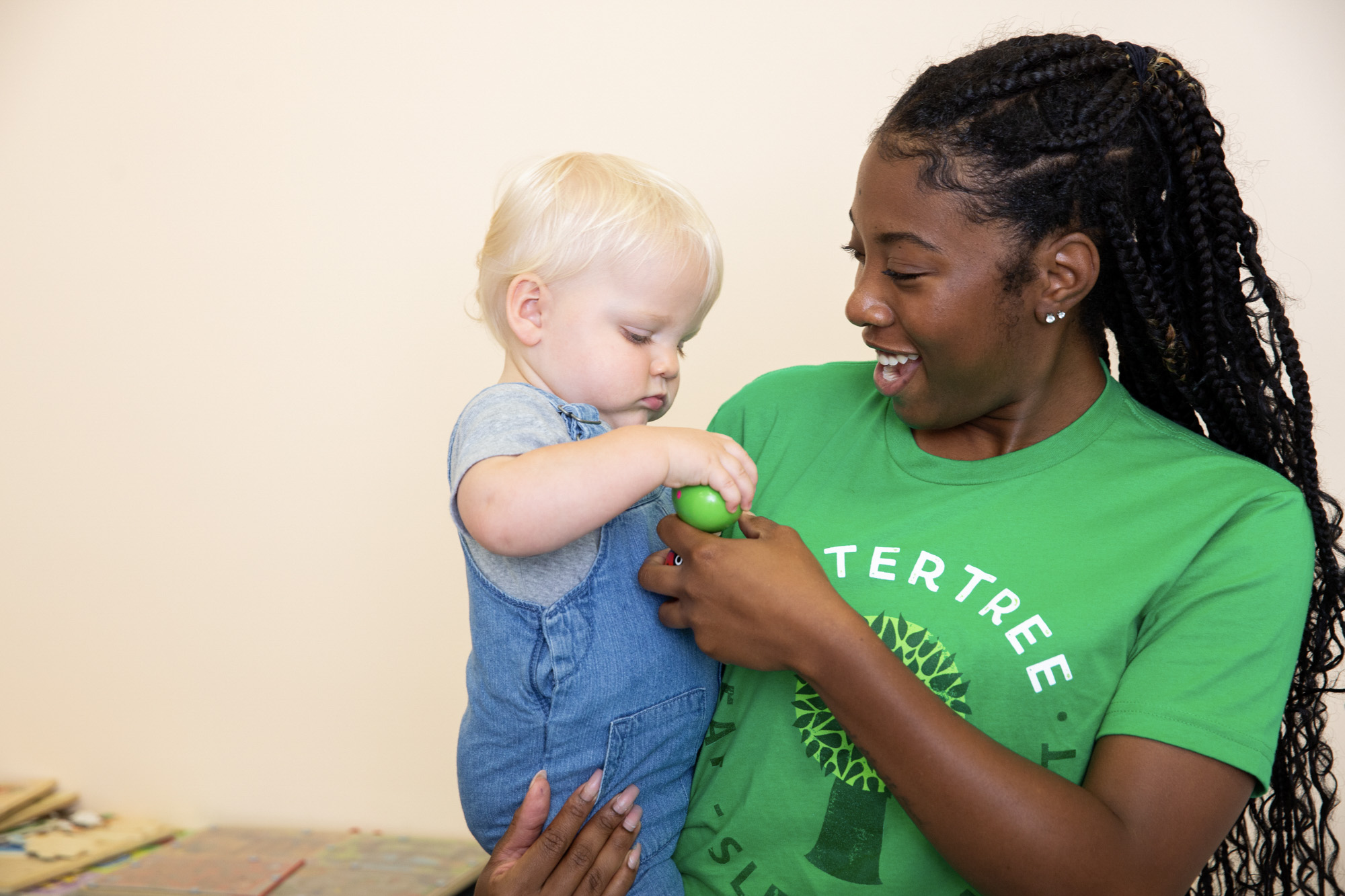 A babysitter wearing a green SitterTree t-shirt holds a toddler dressed in denim overalls, engaging with the child as they explore a small green toy together: Nanny cost vs daycare