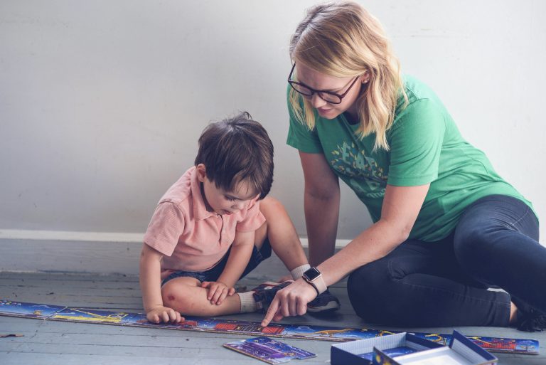 Babysitter with blonde hair and glasses sitting on the floor with a young boy, engaging in a puzzle together, creating a fun and interactive moment: creative babysitting activities