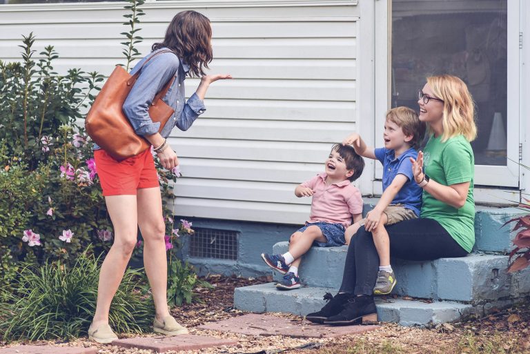 Babysitter sitting on the front steps with two smiling children, waving goodbye to their mother, who is playfully blowing a kiss as she walks away, surrounded by flowers and greenery: what to expect on the first day with a new babysitter