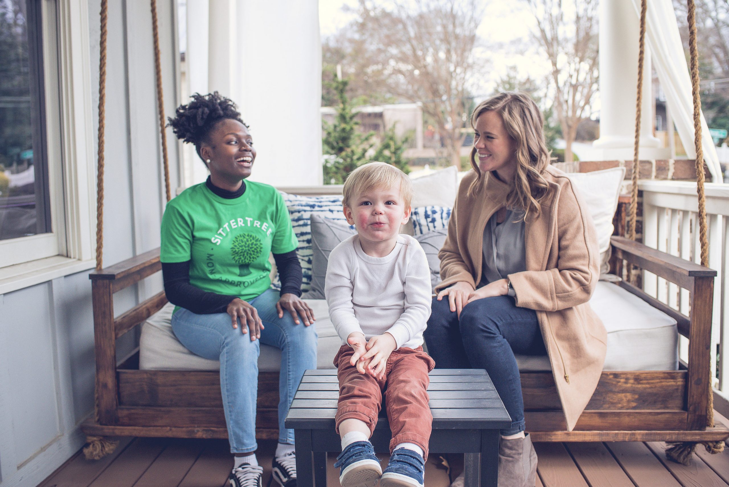 Babysitter and a parent sitting on a cozy porch swing, smiling and chatting, while a young boy sits in the foreground on a small table, looking at the camera with a playful expression: how to address performance issues with your babysitter