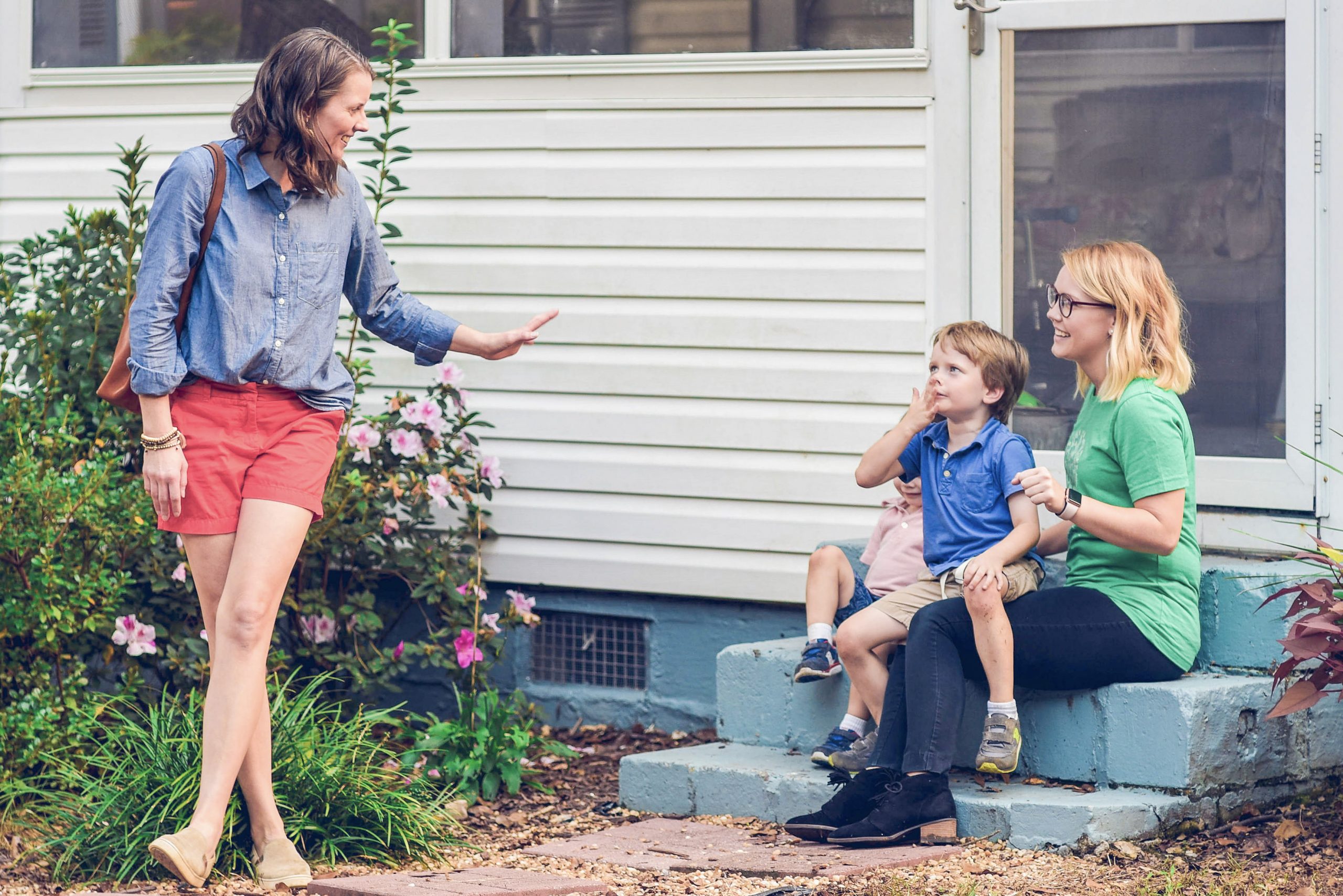 A smiling woman waves goodbye to a babysitter in a green SitterTree T-shirt, who is sitting on outdoor steps with two young children: How to connect with kids and parents