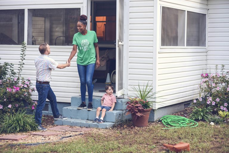 Babysitter greeting a parent with a handshake on the front steps of a house, while a young boy sits nearby, surrounded by a garden with blooming flowers: skills every babysitter should develop