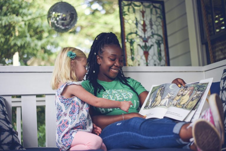 Smiling babysitter with long braided hair reading a picture book with a young blonde girl, sitting on a cozy porch swing surrounded by greenery and stained glass decor: How to handle tantrums as a babysitter