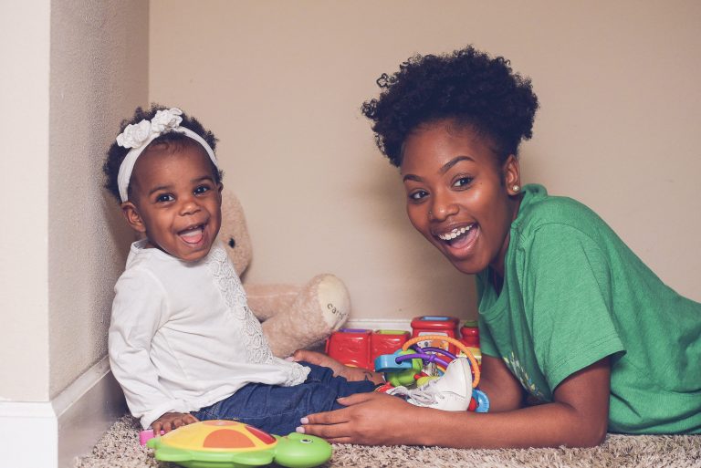 A smiling woman in a green SitterTree T-shirt plays with a happy baby surrounded by colorful toys in a cozy indoor setting: Understanding babysitting taxes