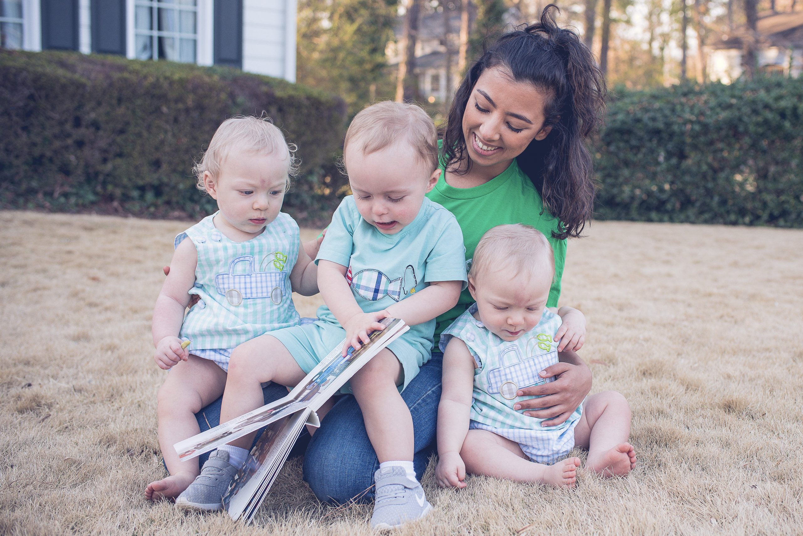 Babysitter in a green SitterTree t-shirt sitting on the grass outdoors, smiling while reading a book to three toddlers dressed in matching outfits, creating a sweet and engaging moment: how to show appreciation to your babysitter