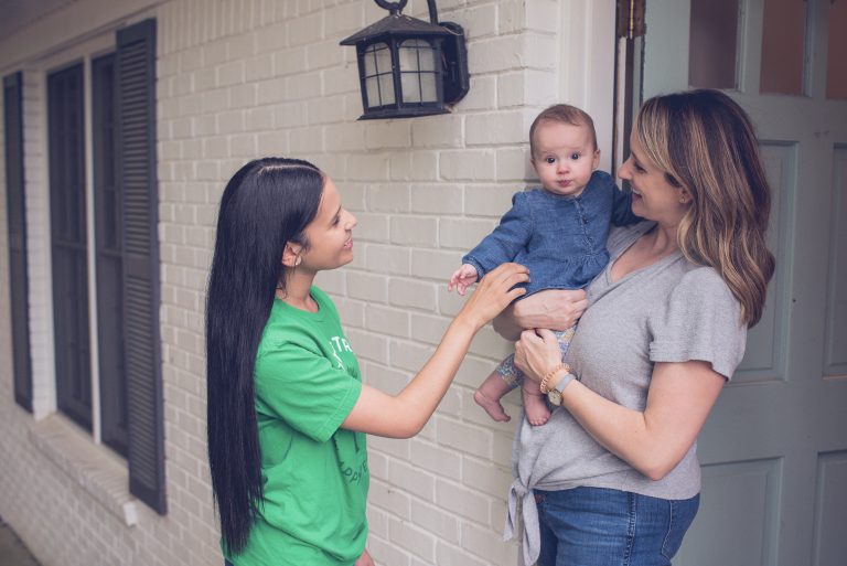 Babysitter with long black hair smiling and greeting a baby being held by their mother at the front door of a brick house, creating a warm and welcoming interaction: how to handle feedback from parents