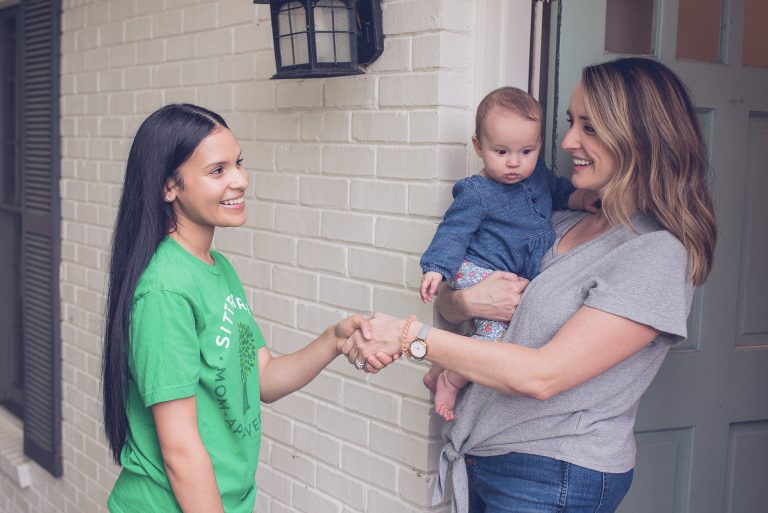 A smiling babysitter wearing a green SitterTree T-shirt greets a mother holding her baby at the front door, shaking hands warmly: How to connect with kids and parents