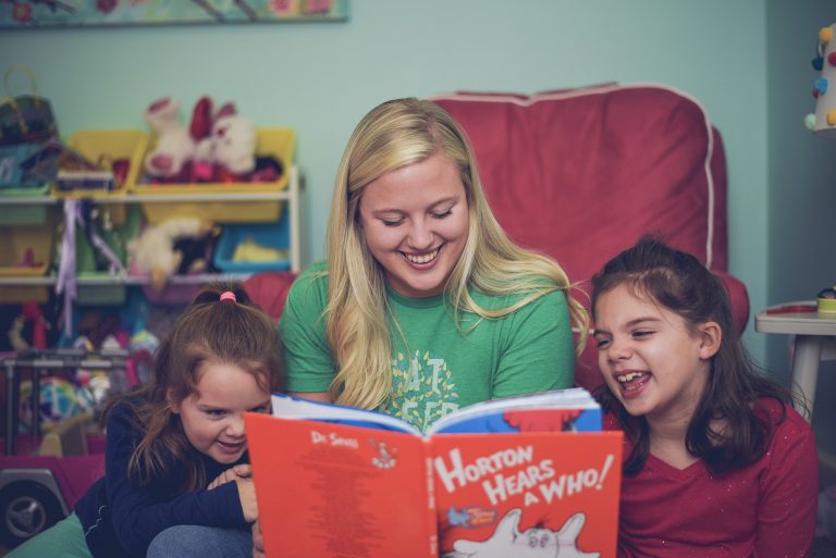 A smiling woman in a green SitterTree T-shirt reads 'Horton Hears a Who!' to two young girls, who are laughing and engaged, in a colorful playroom: how to get babysitting jobs