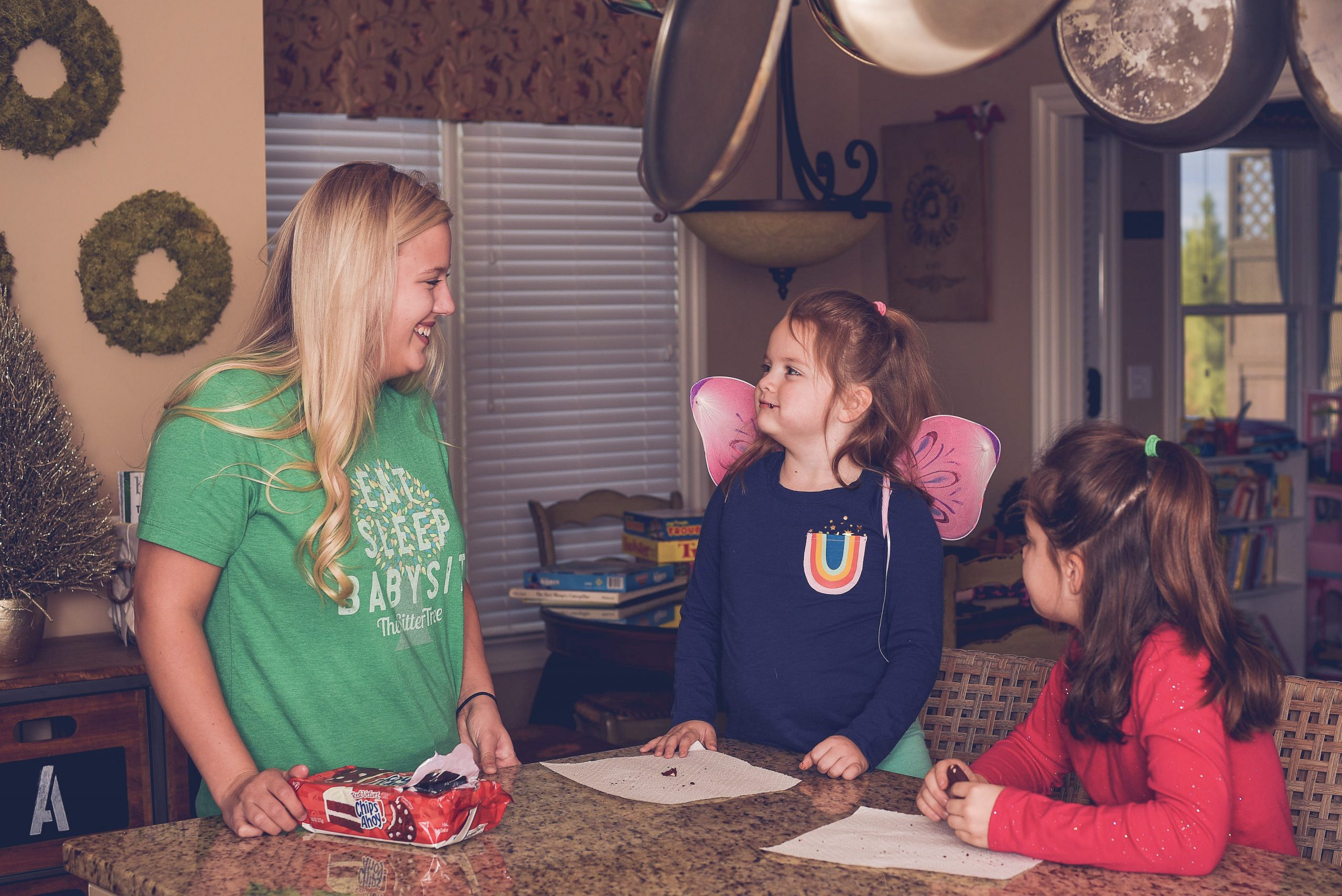 A smiling woman in a green SitterTree T-shirt interacts with two young girls dressed in colorful outfits, standing at a kitchen counter with snacks and paper towels: how to get babysitting jobs