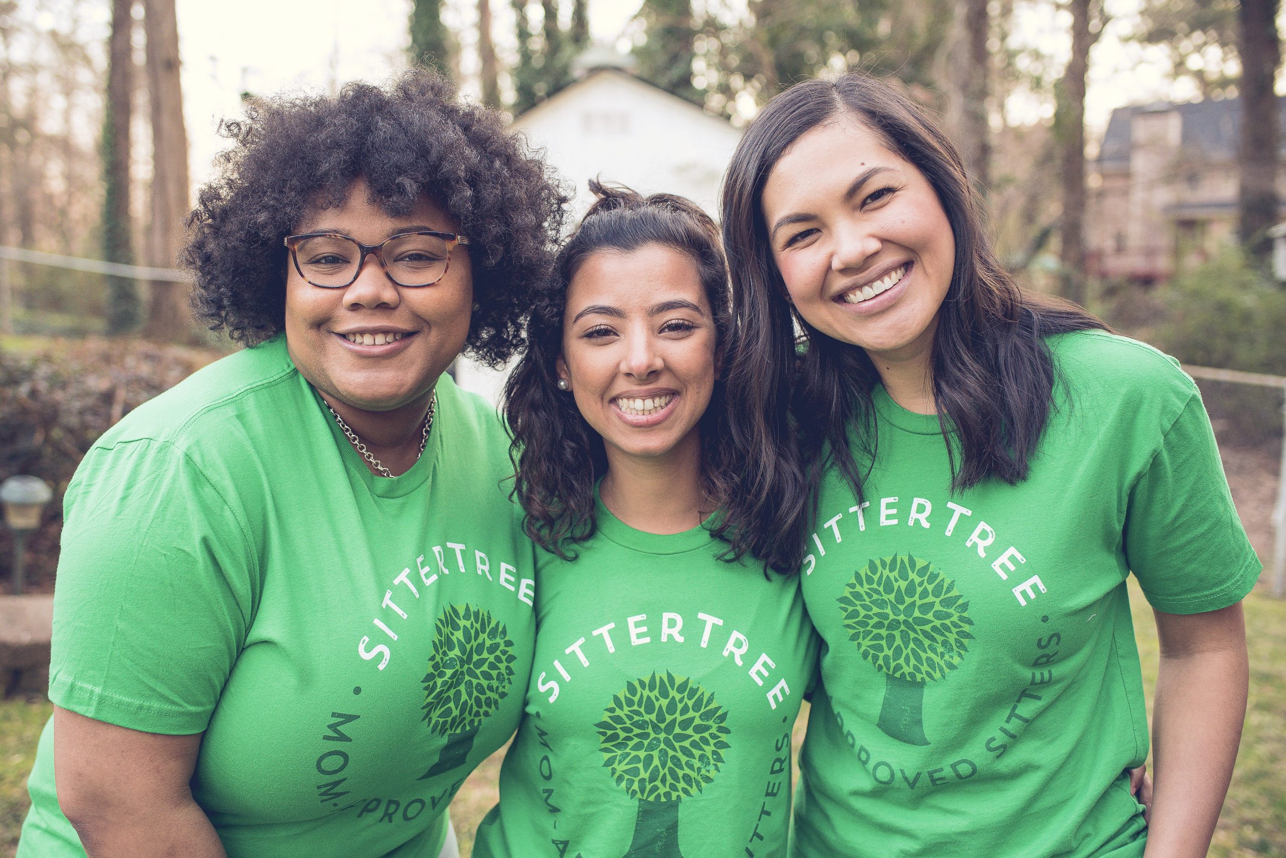 Three smiling babysitters wearing green SitterTree t-shirts standing close together outdoors, showcasing their camaraderie and joyful expressions in a natural setting: Setting Minimum Hours for Babysitting