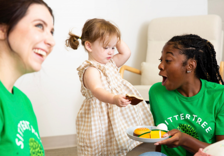 background checks for babysitters: A smiling SitterTree sitter wearing a green SitterTree T-shirt playfully interacting with a toddler holding a toy, creating a fun and engaging moment