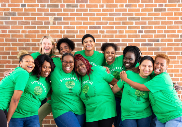 Babysitter vs. Nanny: A group of diverse sitters wearing green SitterTree T-shirts, smiling and embracing each other in front of a brick wall, showcasing a sense of community and teamwork.