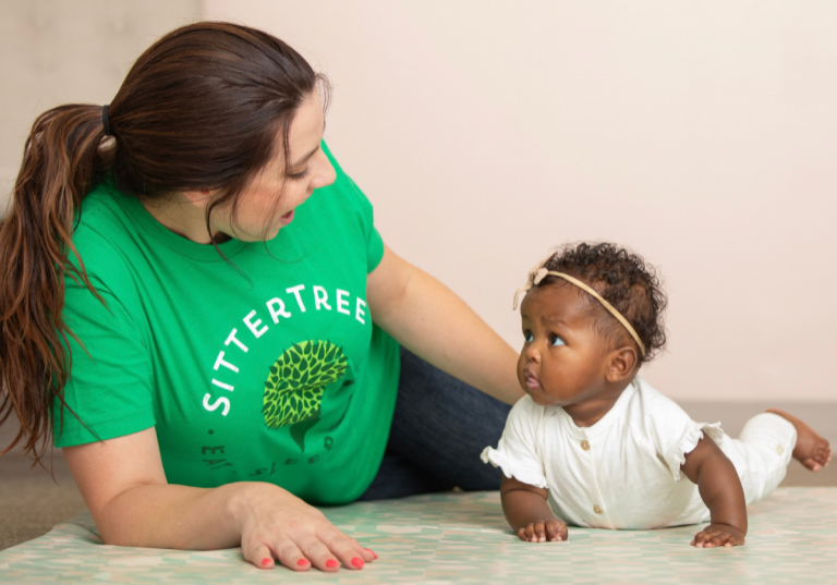 Choosing between a babysitter and a nanny: A SitterTree caregiver lying on the floor engaging with an adorable baby girl, who is looking up with curiosity. The caregiver is wearing a green SitterTree t-shirt, creating a warm and interactive moment