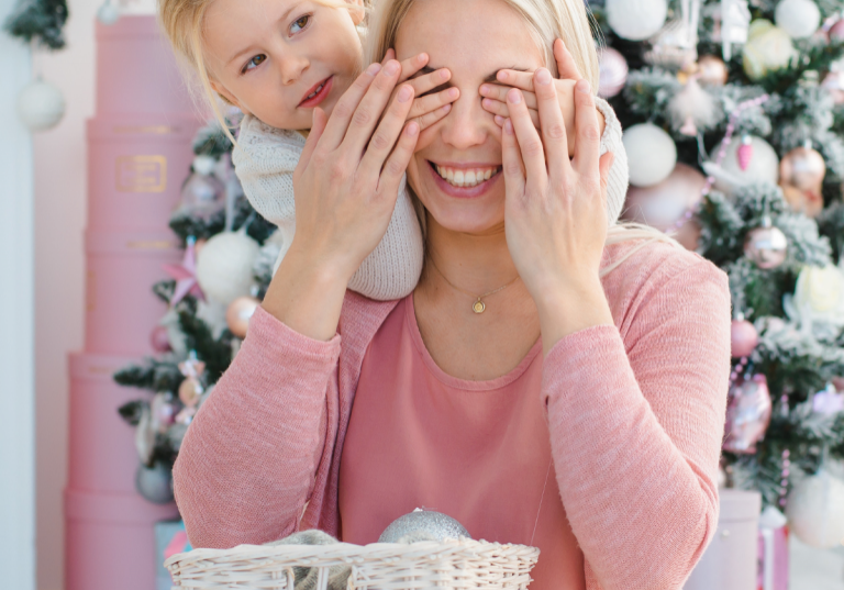 Holiday Gift Guide for Mom: Young child playfully covering their smiling mother's eyes in front of a beautifully decorated Christmas tree, with pink and white holiday decor in the background