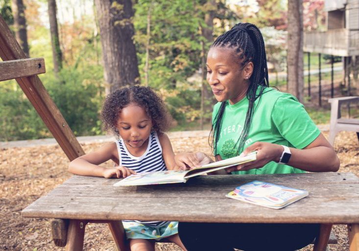 A babysitter wearing a green SitterTree t-shirt sits at an outdoor picnic table with a young girl, reading a colorful book together in a shaded wooded area with a swing set nearby: Nanny cost vs daycare