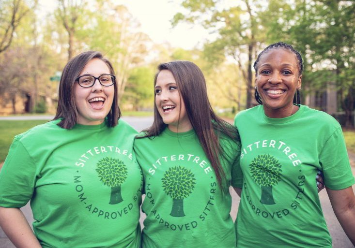 Three cheerful babysitters wearing matching green SitterTree t-shirts, standing outdoors in a sunlit, leafy neighborhood, smiling and enjoying each other's company: Find a nanny near me