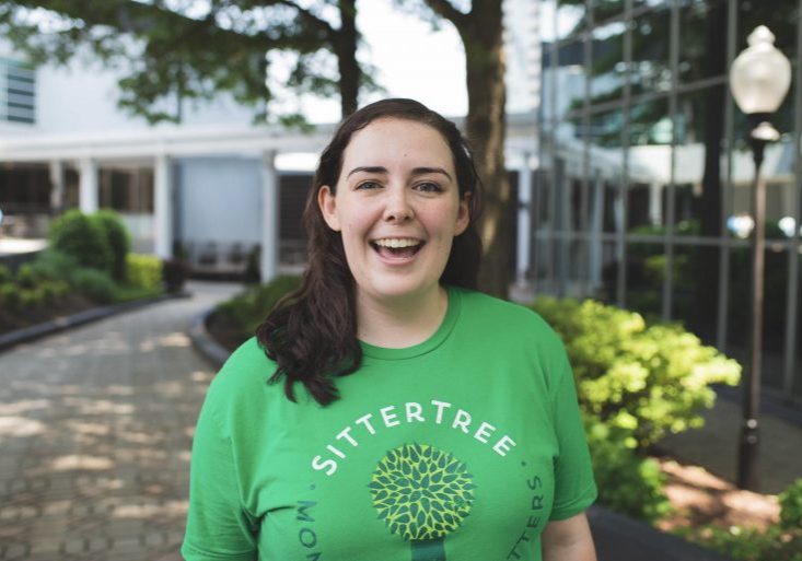 "Smiling babysitter wearing a green SitterTree t-shirt, standing outdoors in a landscaped area with trees and modern buildings in the background: how to verify babysitter references