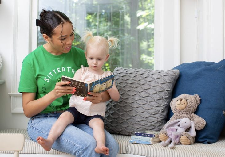 "A babysitter in a green SitterTree t-shirt sitting on a cushioned bench, reading a children's book to a young girl with blonde pigtails, surrounded by cozy pillows and stuffed animals in a bright and inviting room: How to find a nanny