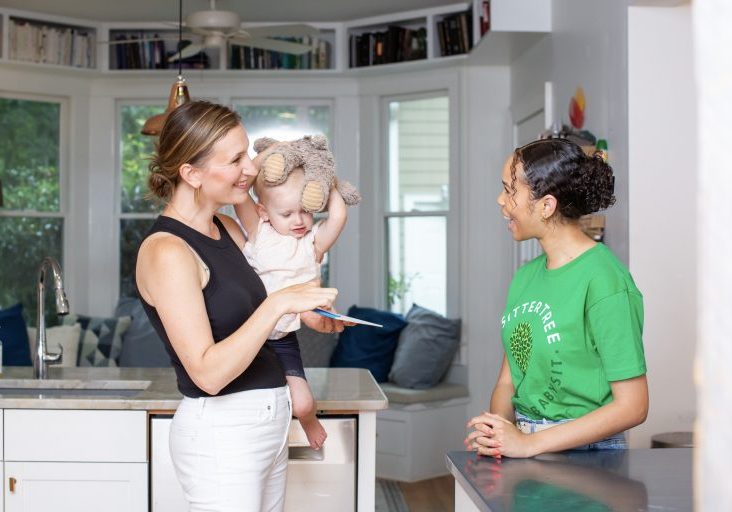 Questions to ask a nanny in an interview: A SitterTree caregiver chatting with a smiling mother holding her young child in a kitchen. The child holds a stuffed animal, and everyone appears engaged and happy in the welcoming home environment