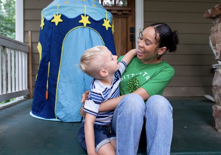 Questions to ask a babysitter: A SitterTree caregiver sitting on a porch, smiling and interacting with a young boy in a striped shirt. They share a joyful moment as the boy leans in with his arm around her, with a play tent visible in the background