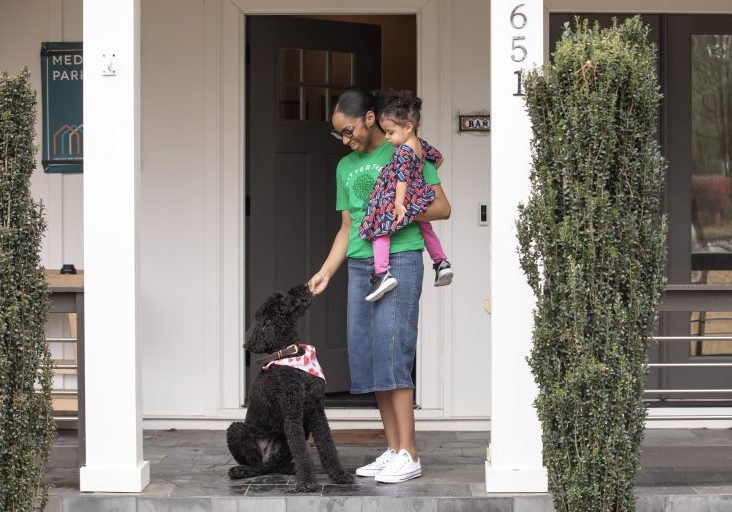 A babysitter wearing a green SitterTree t-shirt stands on a porch holding a toddler in colorful clothing, smiling as they interact with a black poodle wearing a bandana: How to find a babysitter near me