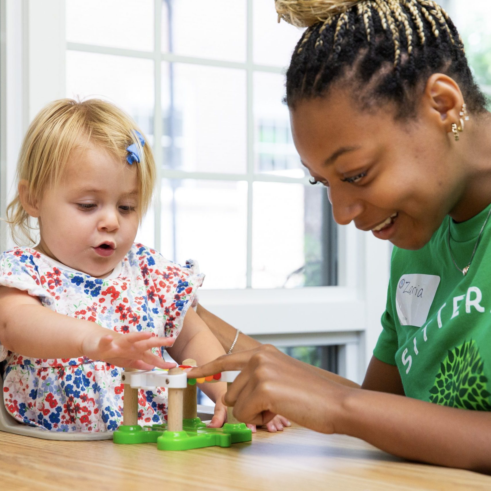 A young woman wearing a green SitterTree t-shirt interacts with a toddler playing with a toy at a wooden table in a brightly lit room: Best babysitting apps and websites