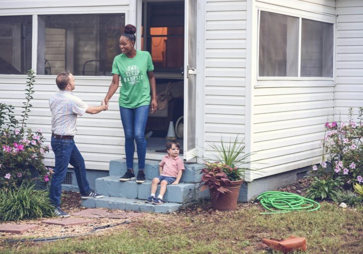 Babysitter greeting a parent with a handshake on the front steps of a house, while a young boy sits nearby, surrounded by a garden with blooming flowers: skills every babysitter should develop