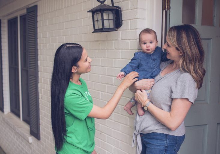 Babysitter with long black hair smiling and greeting a baby being held by their mother at the front door of a brick house, creating a warm and welcoming interaction: how to handle feedback from parents